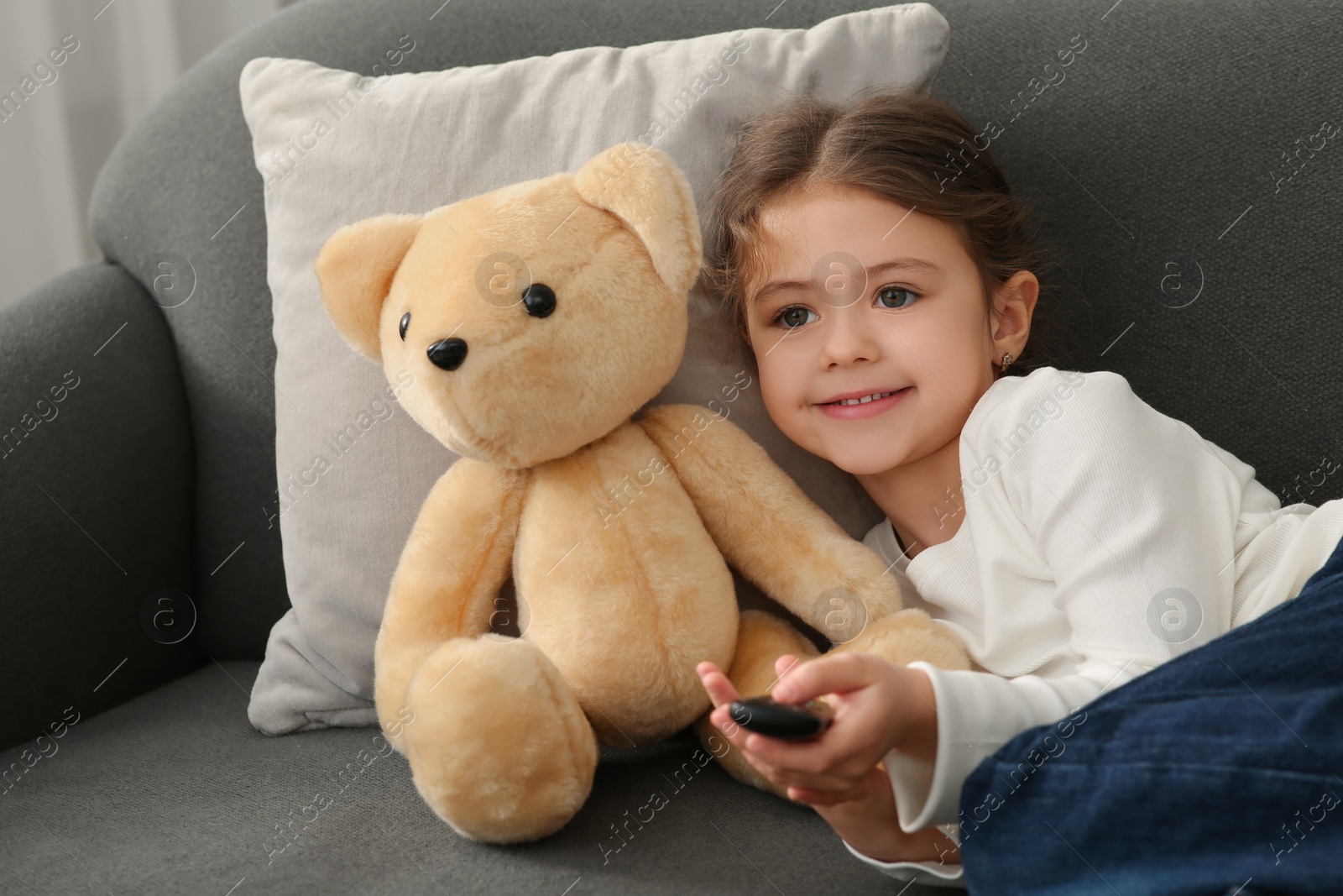 Photo of Little girl with toy bear changing TV channels by remote control on sofa at home