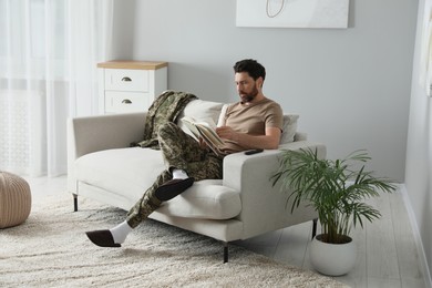 Photo of Soldier reading book on soft sofa in living room. Military service