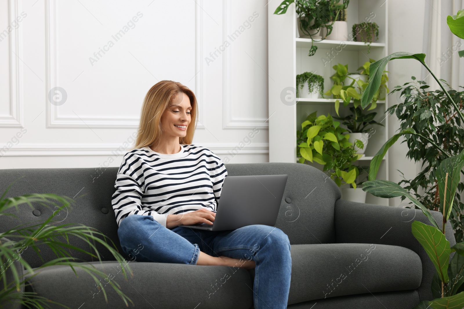 Photo of Woman working with laptop on sofa surrounded by beautiful potted houseplants at home