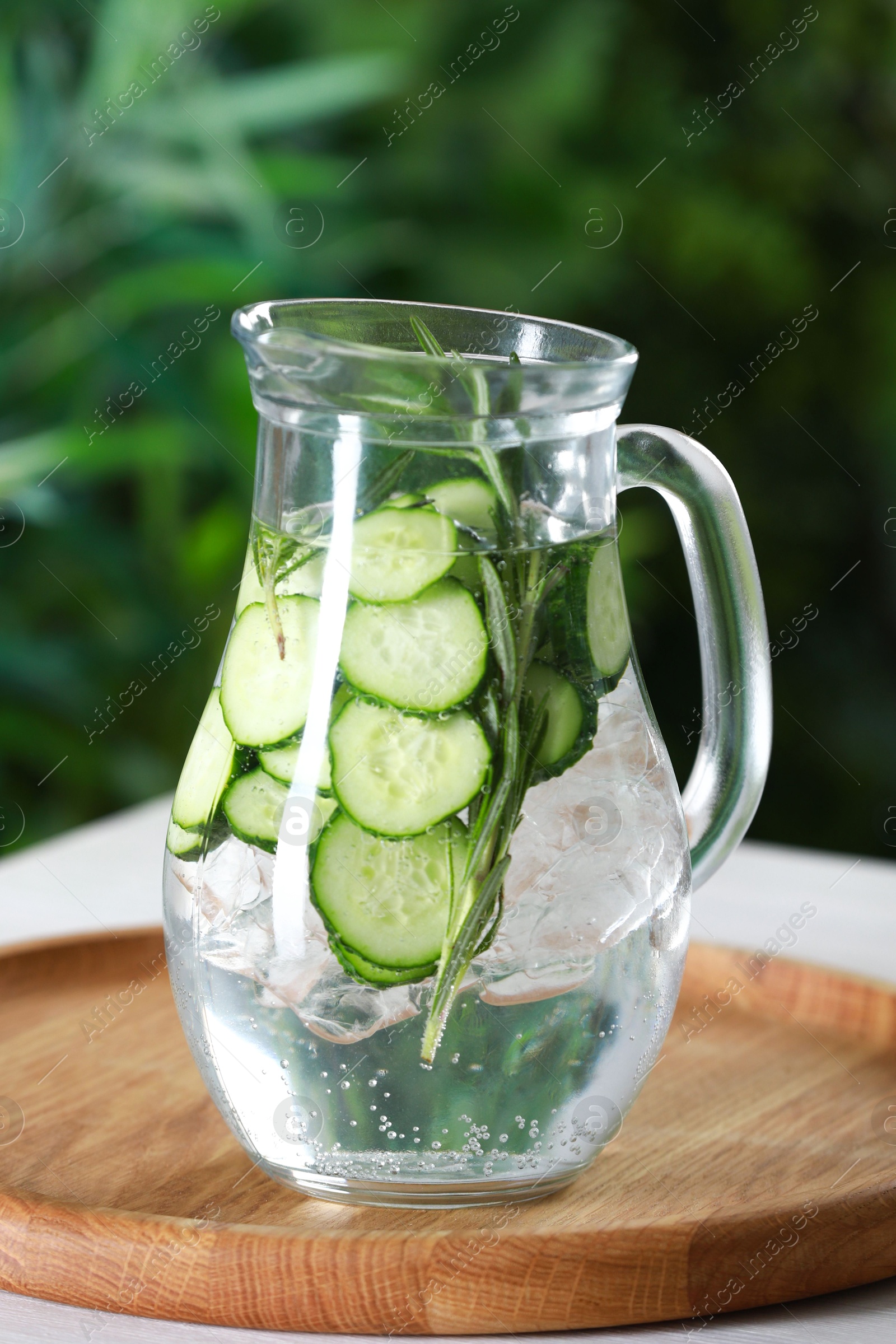 Photo of Refreshing cucumber water with rosemary in jug on table against blurred green background