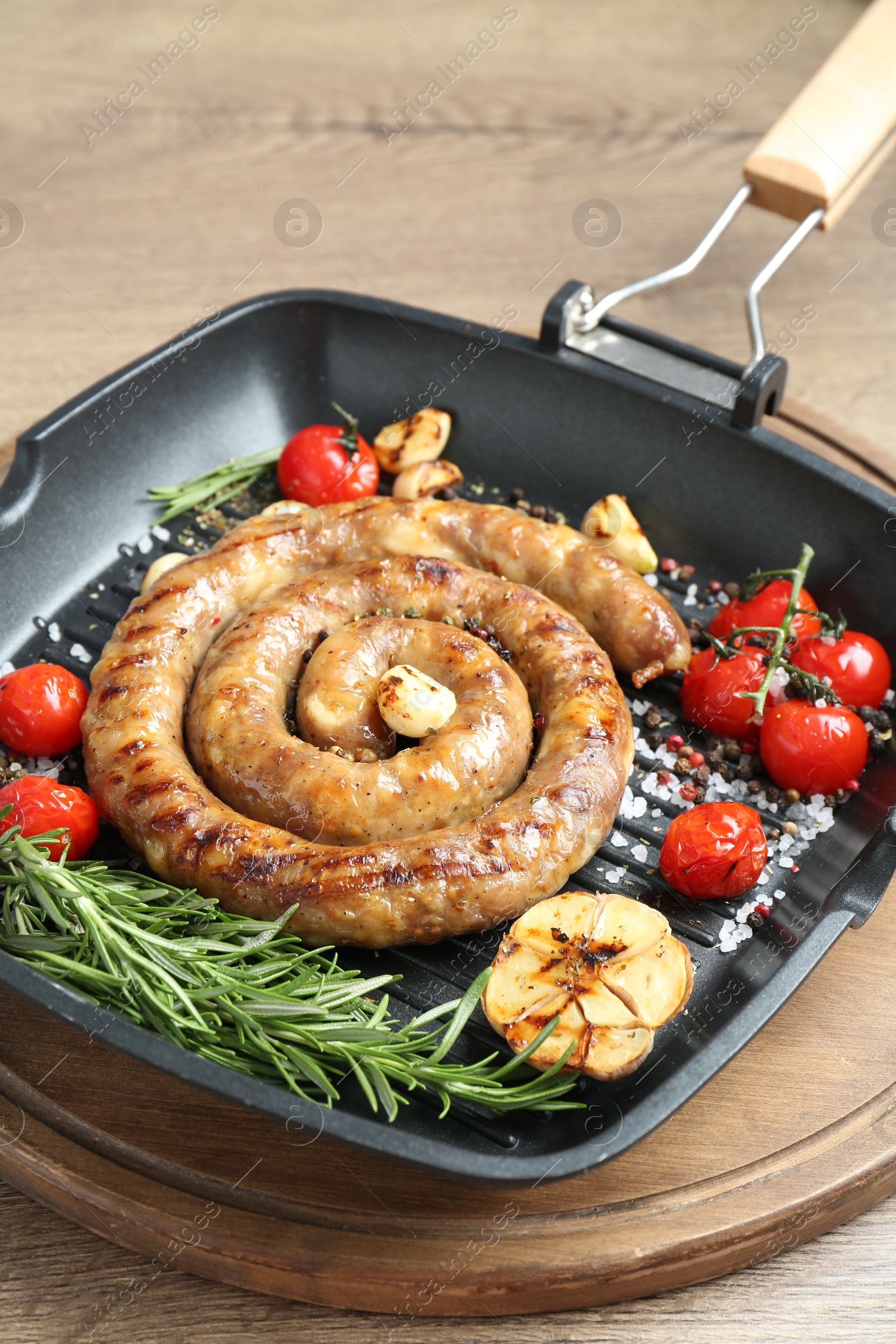Photo of Delicious homemade sausage with garlic, tomatoes, rosemary and spices in grill pan on wooden table, closeup
