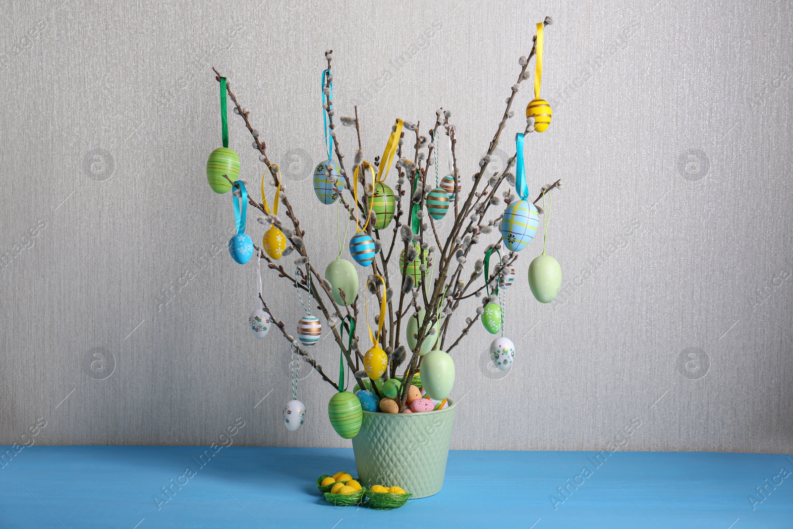 Photo of Beautiful willow branches with painted eggs in vase on light blue table. Easter decor