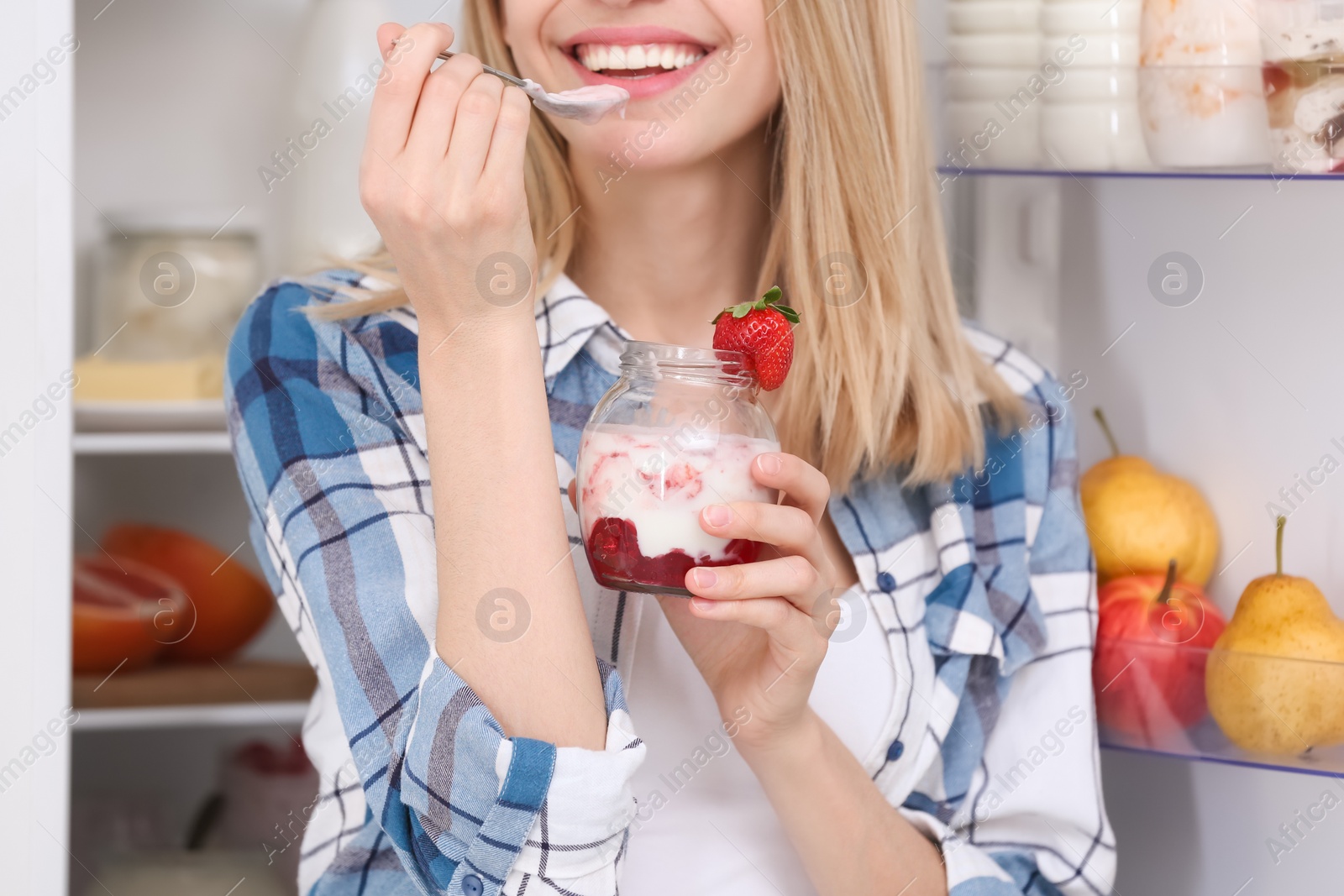 Photo of Young attractive woman eating tasty yogurt near fridge