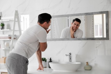 Handsome man touching his smooth face after shaving near mirror in bathroom