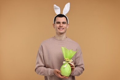 Photo of Easter celebration. Handsome young man with bunny ears holding wrapped gift on beige background