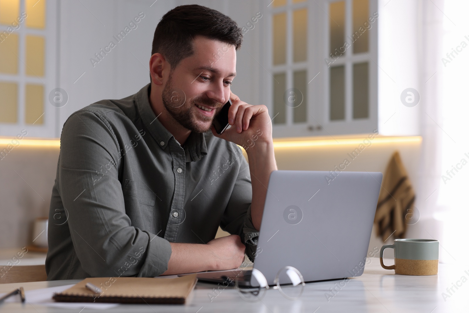 Photo of Happy man talking on phone while working with laptop at white table in kitchen