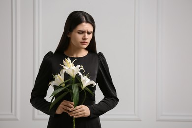 Sad woman with lilies mourning near white wall, space for text. Funeral ceremony