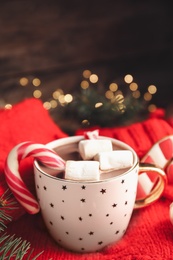 Photo of Cup of tasty cocoa with marshmallows and Christmas candy cane on knitted sweater against blurred festive lights. Space for text