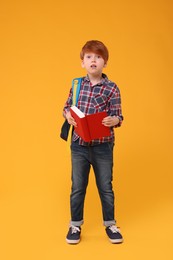 Cute schoolboy with backpack and book on orange background