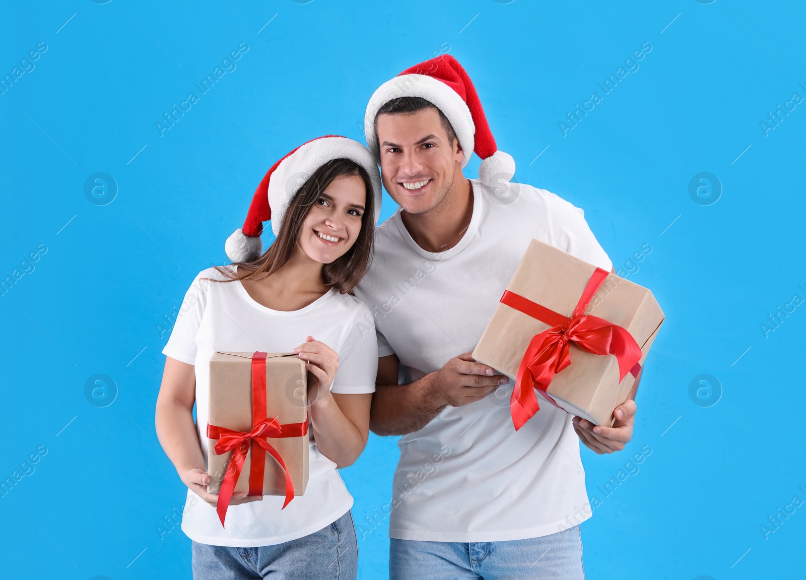 Photo of Beautiful happy couple in Santa hats holding Christmas gifts on light blue background