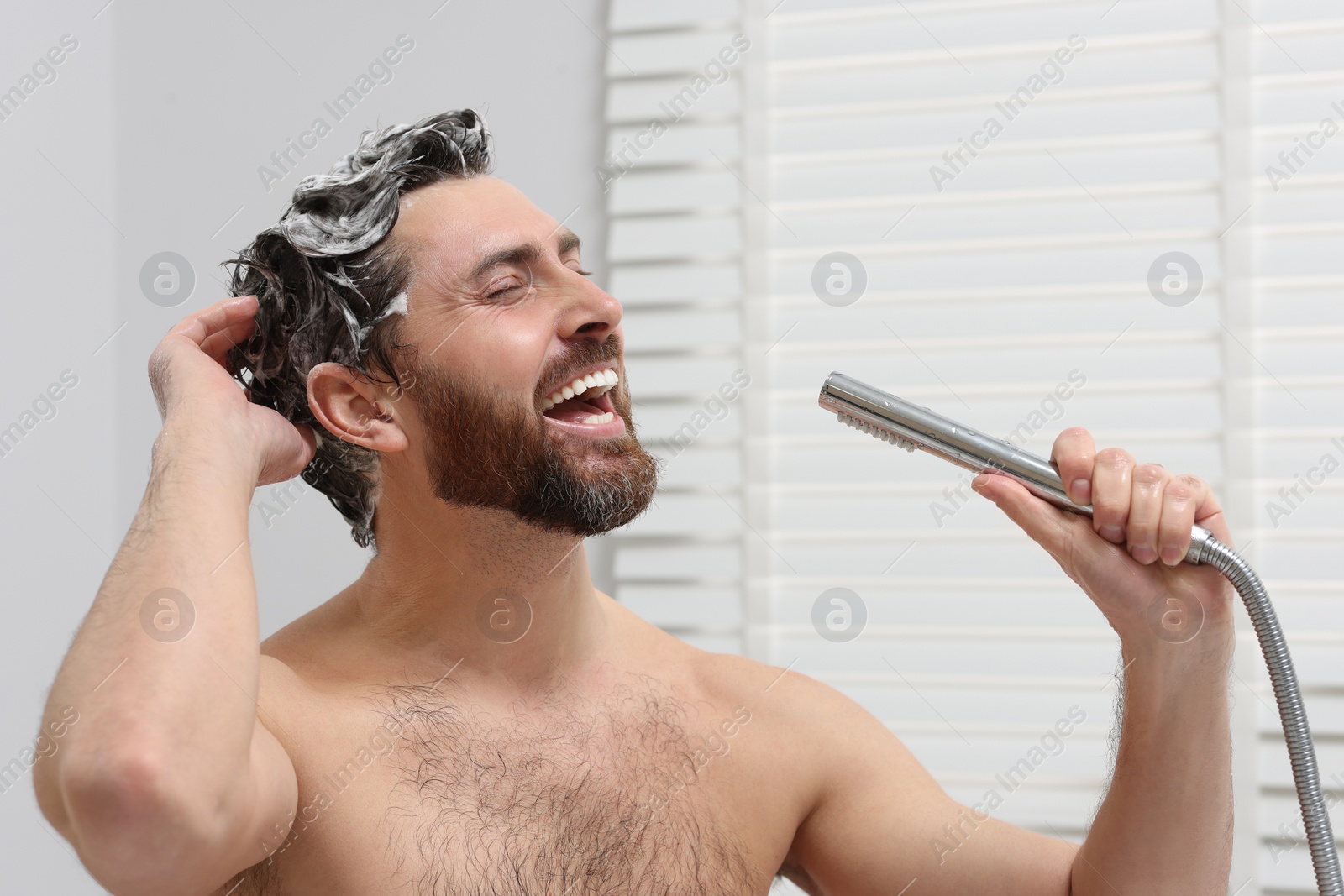 Photo of Happy man with showerhead singing and washing his hair with shampoo indoors