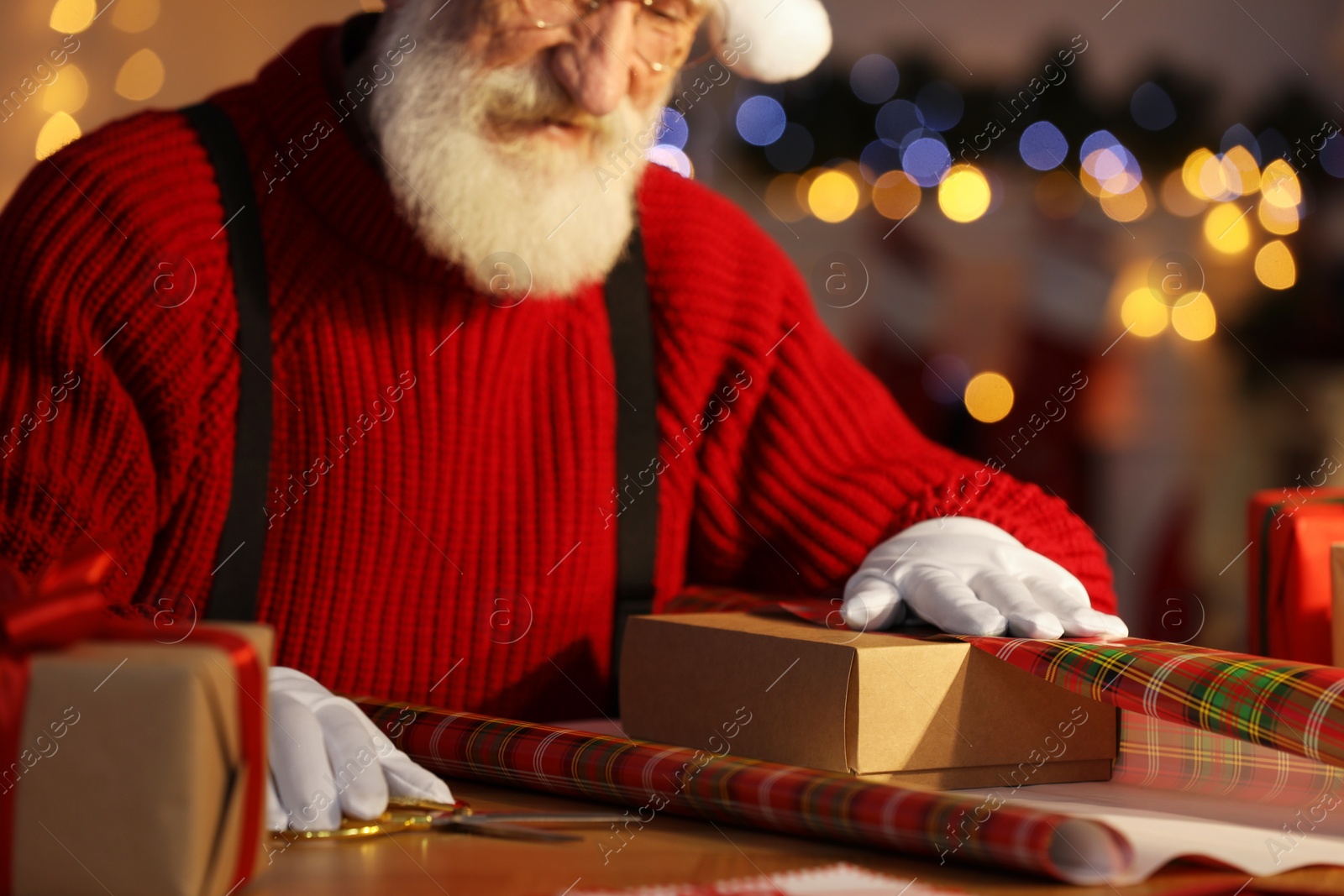 Photo of Santa Claus wrapping gift at his workplace in room decorated for Christmas, closeup