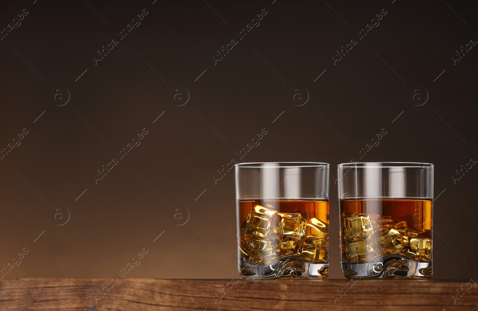 Photo of Whiskey with ice cubes in glasses on wooden table against brown background, space for text