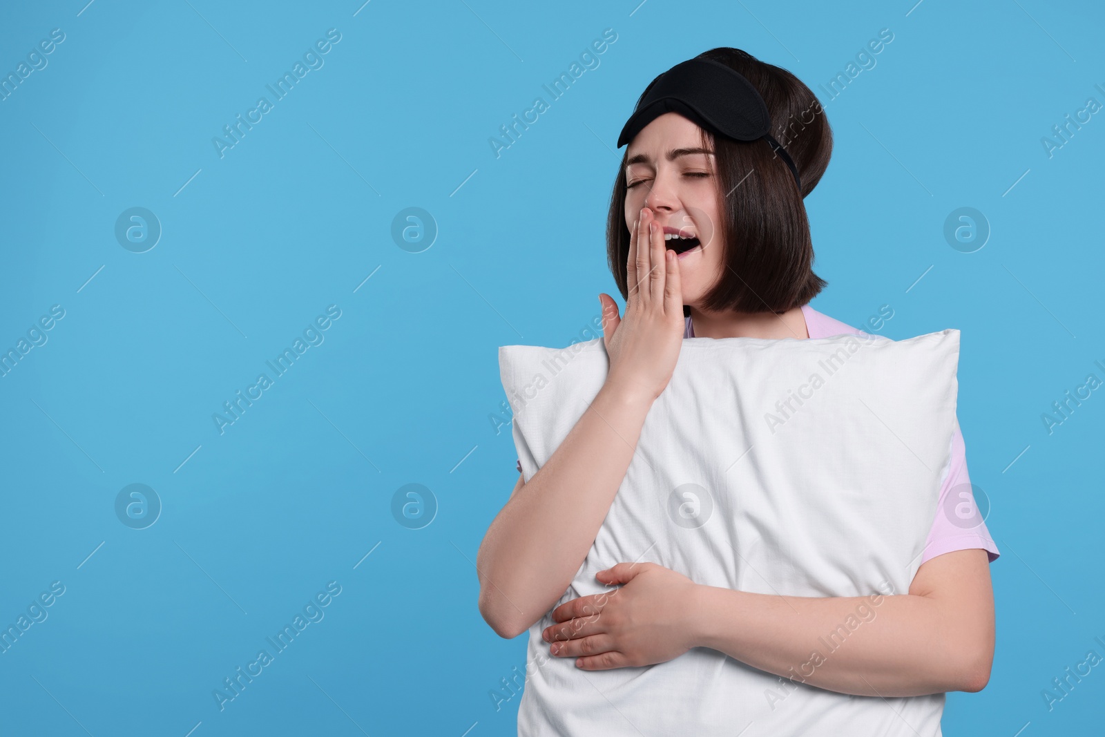 Photo of Sleepy young woman with sleep mask and pillow yawning on light blue background, space for text. Insomnia problem