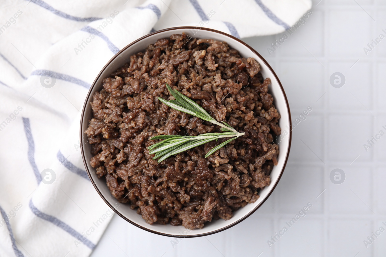 Photo of Fried ground meat in bowl and rosemary on white tiled table, top view