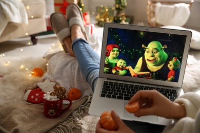 Photo of MYKOLAIV, UKRAINE - DECEMBER 25, 2020: Woman with tangerine watching Shrek The Halls movie on laptop at home, closeup. Cozy winter holidays atmosphere
