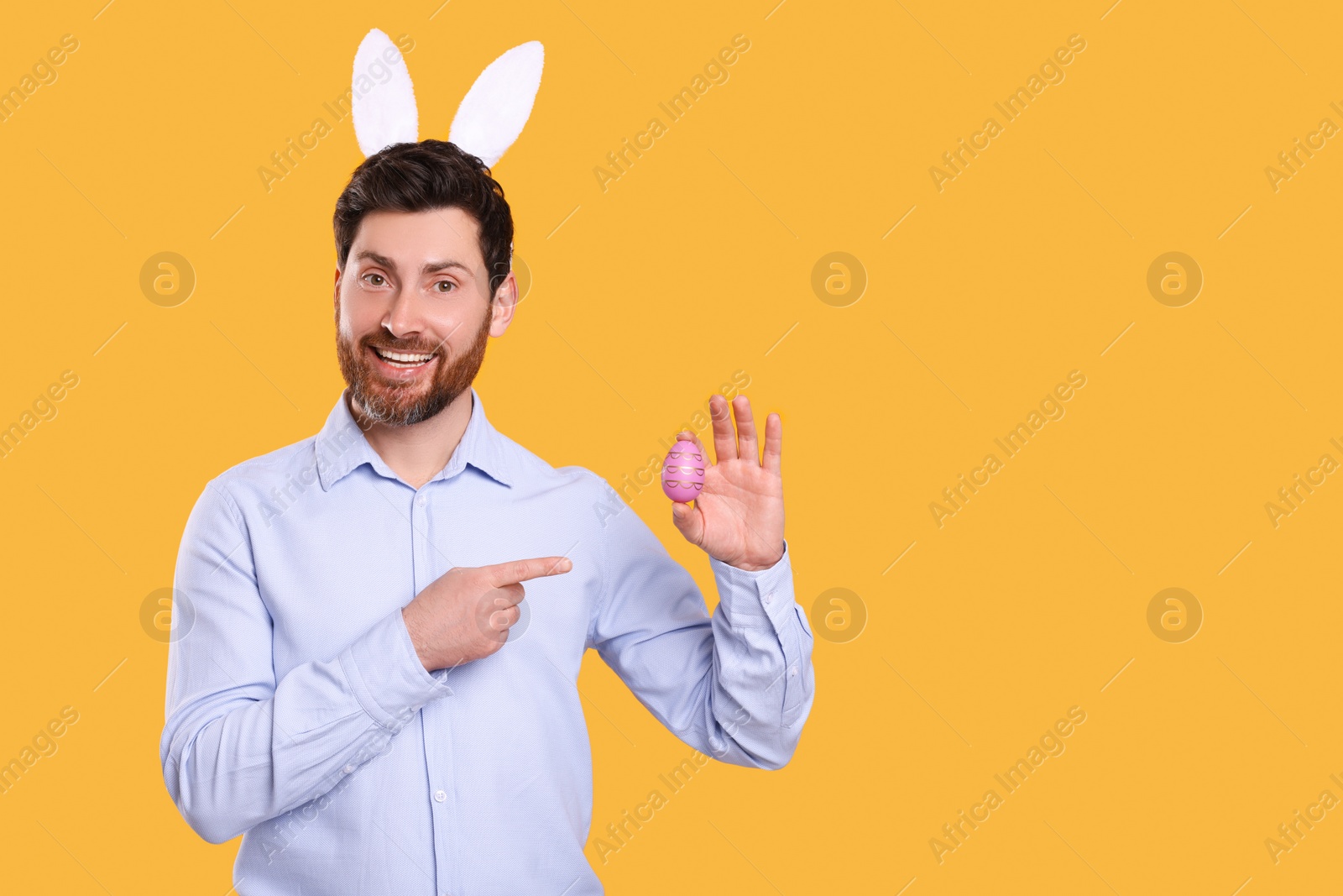 Photo of Portrait of happy man in cute bunny ears headband pointing at Easter egg on orange background. Space for text