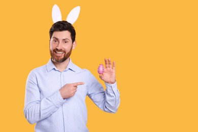 Portrait of happy man in cute bunny ears headband pointing at Easter egg on orange background. Space for text
