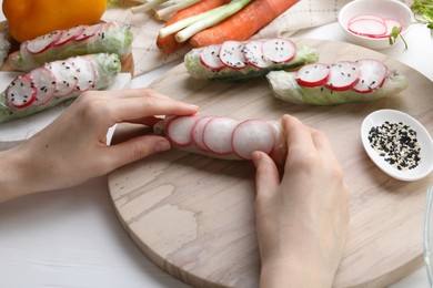 Photo of Woman wrapping spring roll at white wooden table with products, closeup