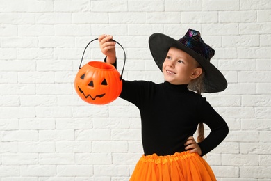 Photo of Cute little girl with pumpkin candy bucket wearing Halloween costume near white brick wall