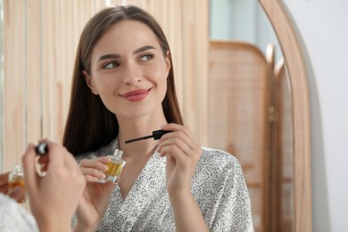 Young woman with eyelash oil near mirror indoors, space for text