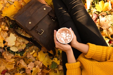 Photo of Woman holding cup of hot drink in park with fallen leaves, above view. Autumn season