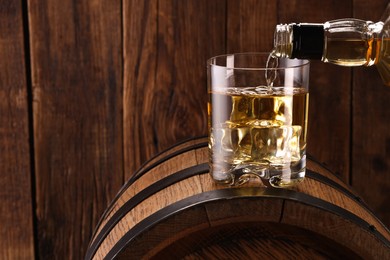 Photo of Pouring whiskey from bottle into glass on barrel against wooden background, closeup. Space for text