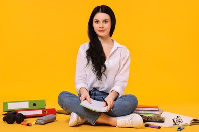 Student with notebook sitting among books and stationery on yellow background