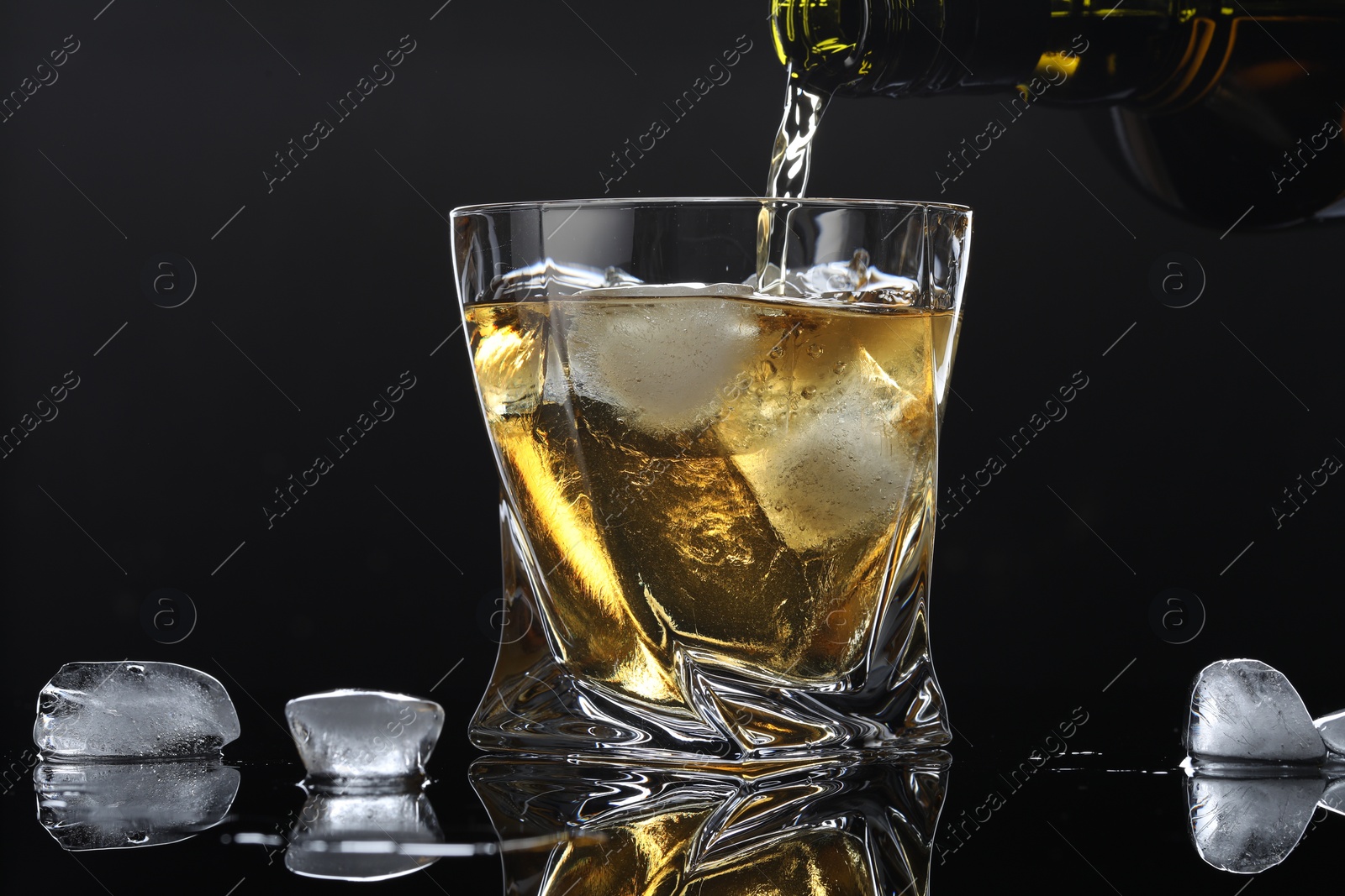 Photo of Pouring tasty whiskey from bottle into glass with ice at mirror table against black background, closeup