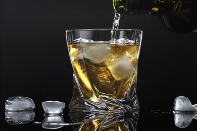 Pouring tasty whiskey from bottle into glass with ice at mirror table against black background, closeup