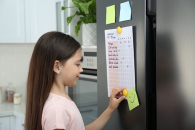 Photo of Little girl putting to do list on fridge in kitchen