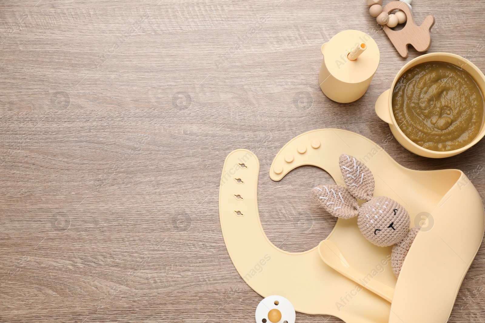 Photo of Silicone baby bib, toys and plastic dishware with healthy food on wooden table, flat lay. Space for text