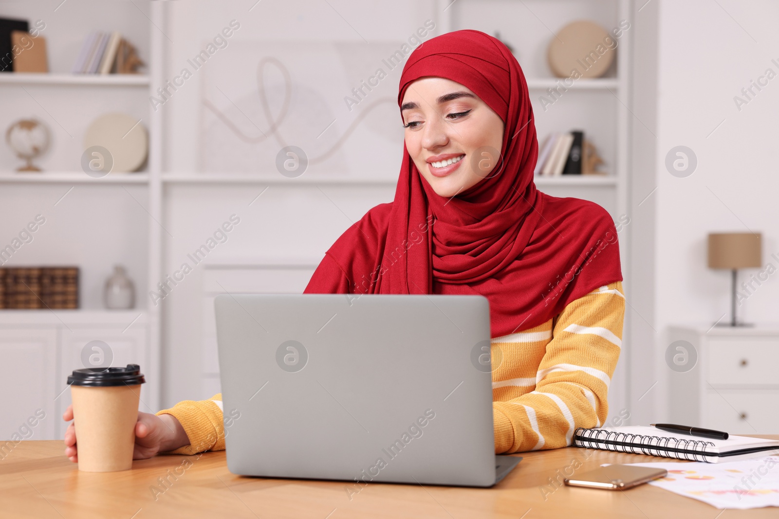 Photo of Muslim woman in hijab using laptop at wooden table in room