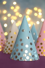 Photo of Beautiful party hats on purple table against blurred lights
