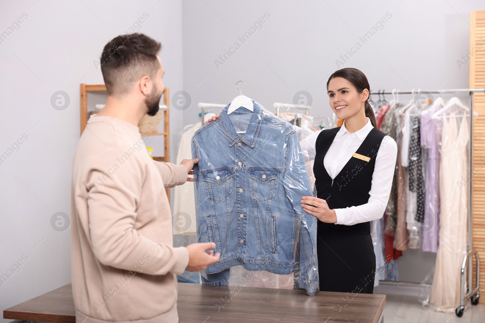 Photo of Dry-cleaning service. Happy worker giving denim jacket in plastic bag to client indoors