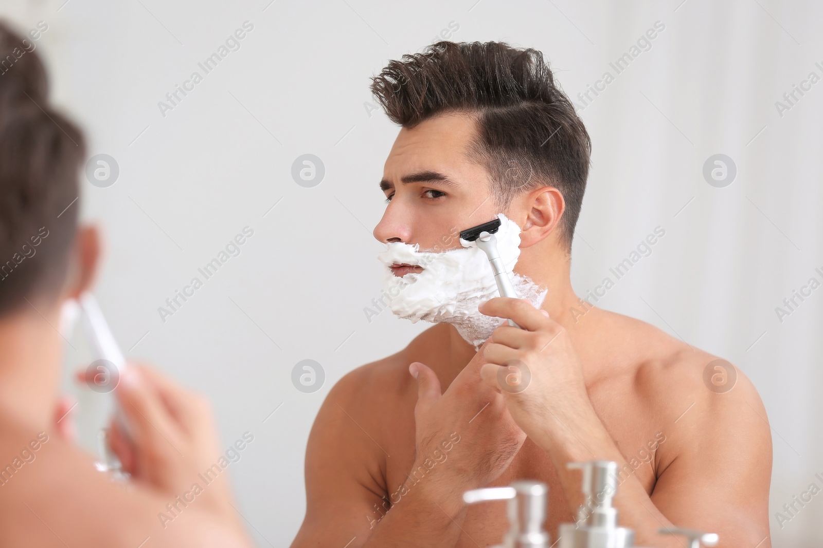 Photo of Young man shaving near mirror in bathroom