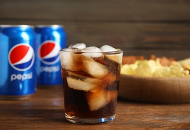 MYKOLAIV, UKRAINE - FEBRUARY 15, 2021: Glass of Pepsi, cans and snack on wooden table