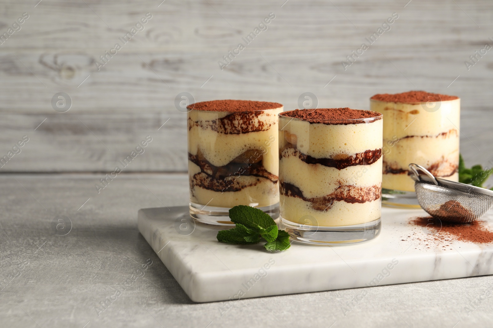 Photo of Delicious tiramisu in glasses, mint and sieve with cocoa powder on grey textured table. Space for text