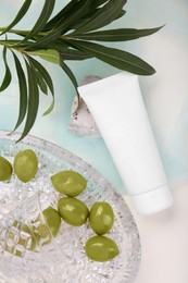 Photo of Tube of cream, olives, stone and leaves on beige background with light blue organza fabric drapery, flat lay