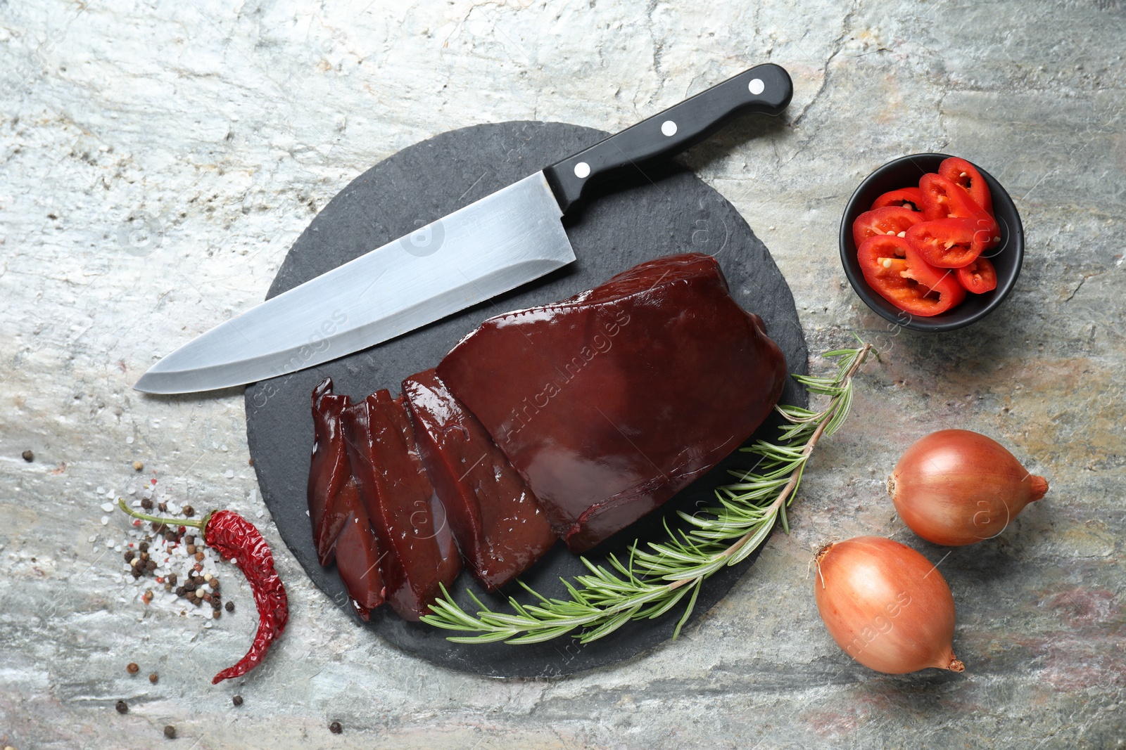 Photo of Cut raw beef liver with rosemary, chili pepper, onions and knife on grey table, flat lay