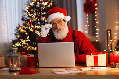 Photo of Santa Claus using laptop and drinking hot beverage at his workplace in room with Christmas tree