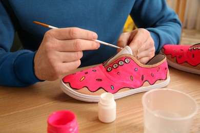 Photo of Man painting on sneaker at wooden table indoors, closeup. Customized shoes