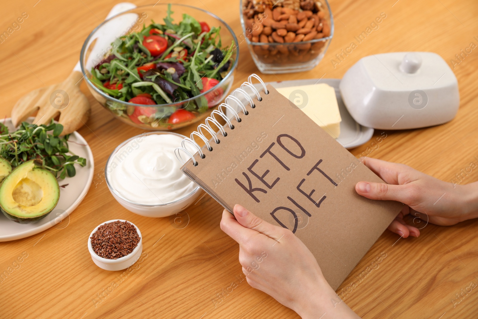 Photo of Woman holding notebook with phrase Keto Diet and different products at wooden table, closeup
