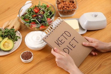 Woman holding notebook with phrase Keto Diet and different products at wooden table, closeup