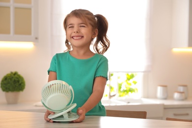 Photo of Little girl enjoying air flow from portable fan at table in kitchen. Summer heat