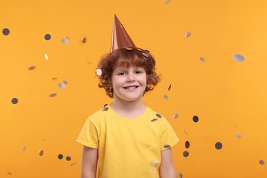 Happy little boy in party hat under falling confetti on orange background