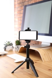 Photo of Tripod with smartphone near computer on table indoors. Mockup for design