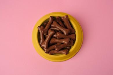 Photo of Yellow bowl with bone shaped dog cookies on pink background, top view