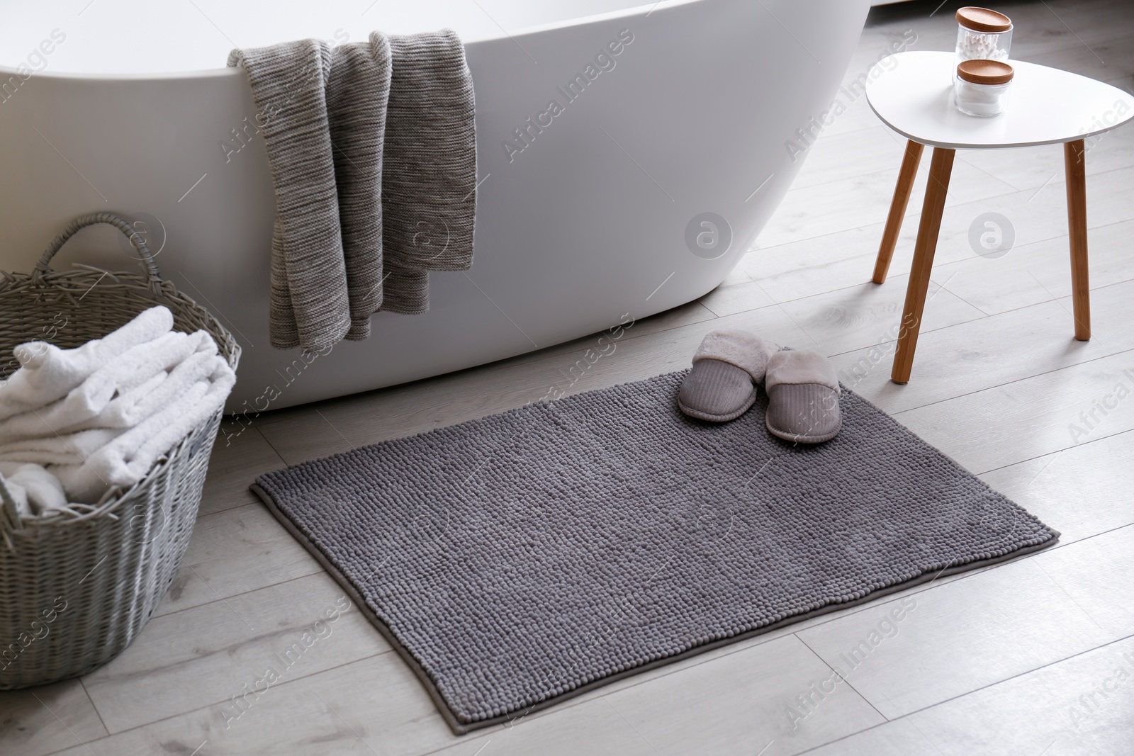 Photo of Soft grey mat with slippers on floor near tub in bathroom. Interior design