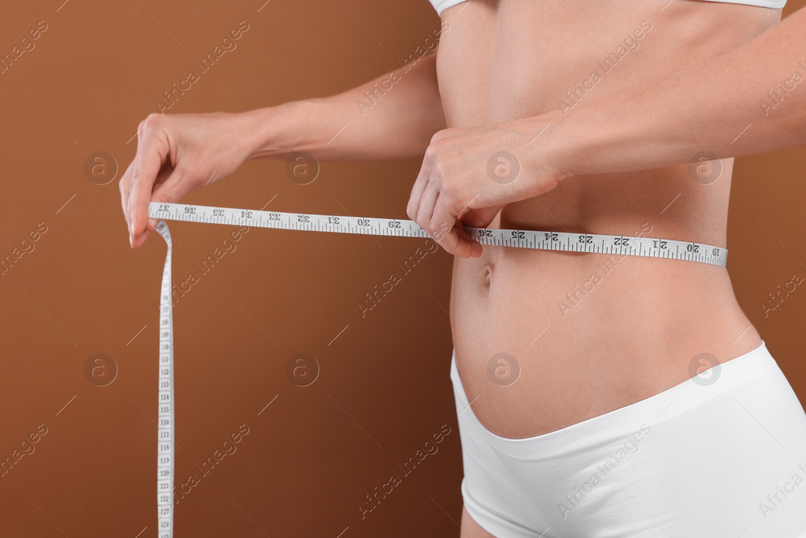 Photo of Slim woman measuring waist with tape on brown background, closeup. Weight loss